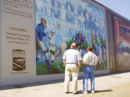 two men look at a section of wall seemingly titled Touchdown, painted with an old football game described in text as the Portsmouth Spartans playing the Green Bay Packers with a score of 19-0